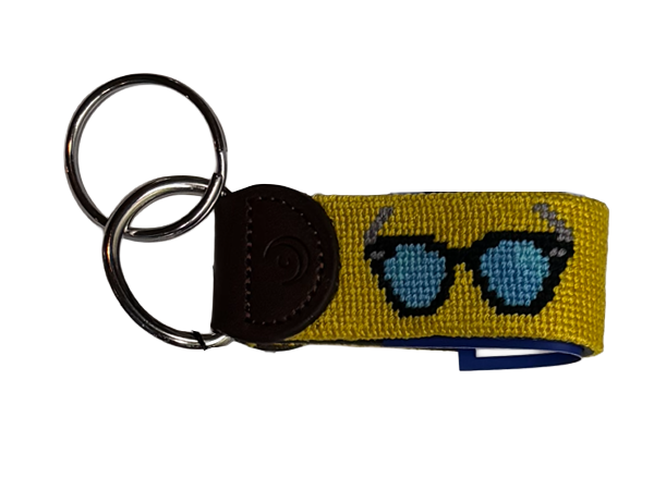A yellow woven keychain strap featuring hand-stitched needlepoint embroidered blue and black sunglasses design, attached to a dark brown leather tab and two silver metal key rings, shown on a plain white background.