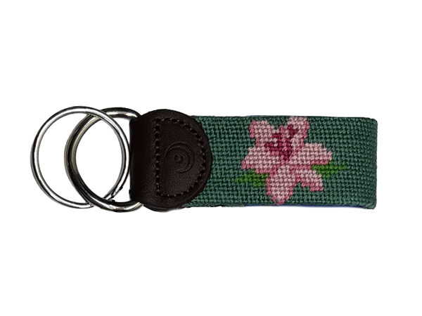 A hand-stitched needlepoint keychain in dark green with a pink embroidered azalea flower, attached to a dark brown leather backing and two stainless steel metal key rings, shown on a plain white background.