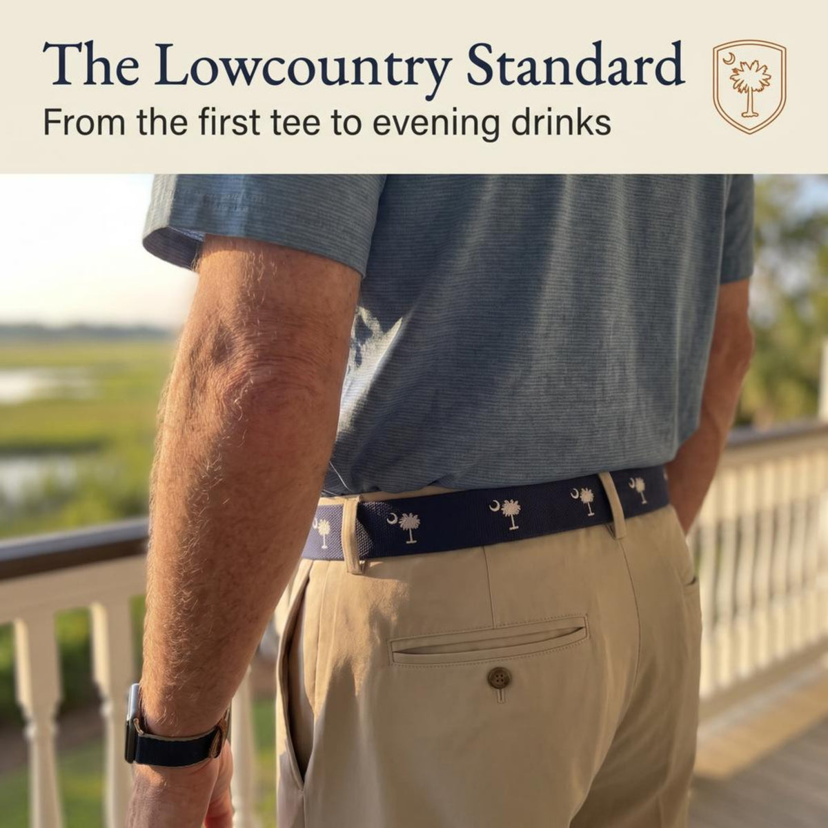 A man wearing a blue short-sleeve polo shirt, khaki pants, and a navy belt with white palm tree motifs, standing on a balcony with a golf course in the background. Text at the top reads 'The Lowcountry Standard' with a small palm tree crest logo. sc flag belt.