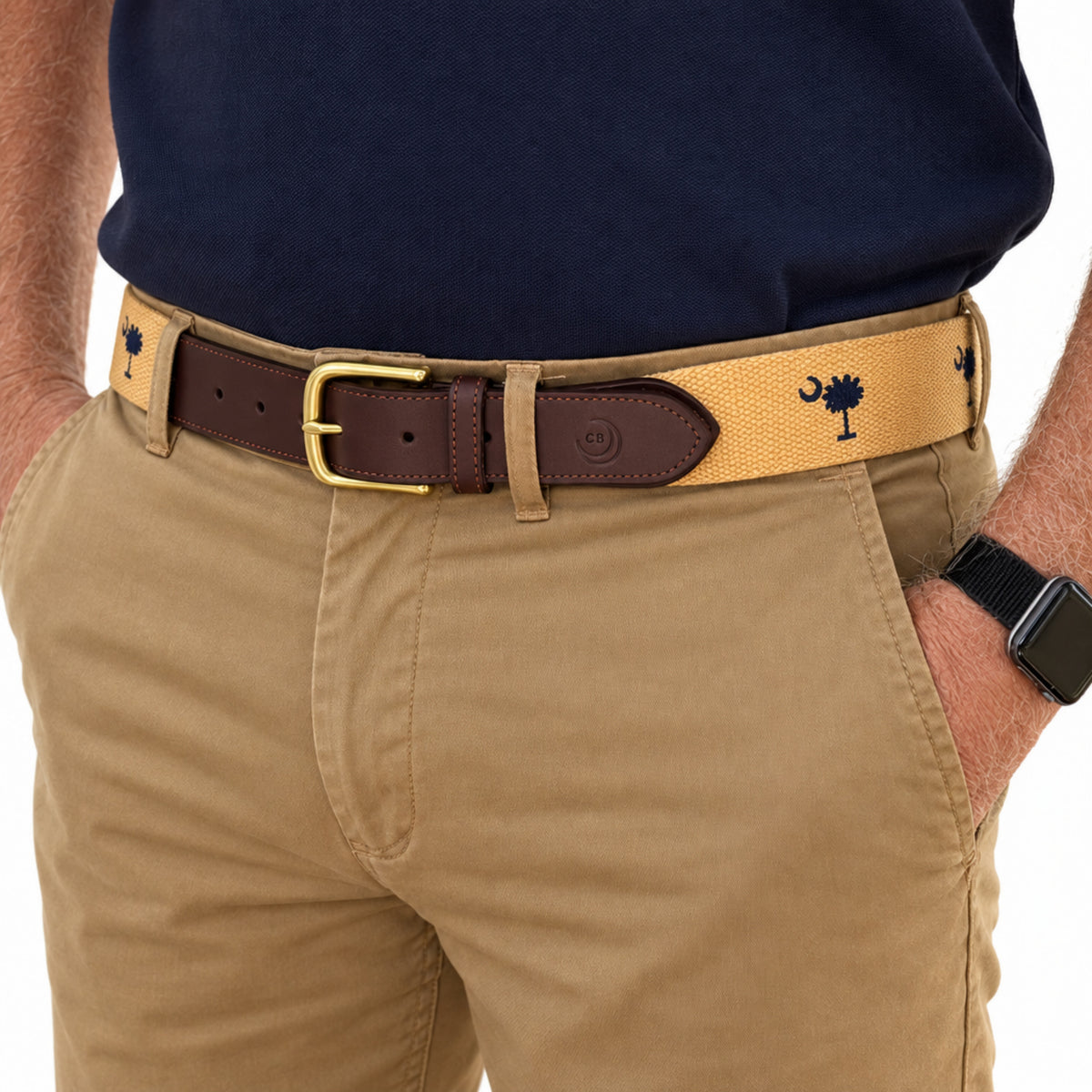 A close-up of a person wearing a tan woven belt with a dark brown leather section and a gold buckle, paired with khaki pants and a navy shirt. sc flag belt.