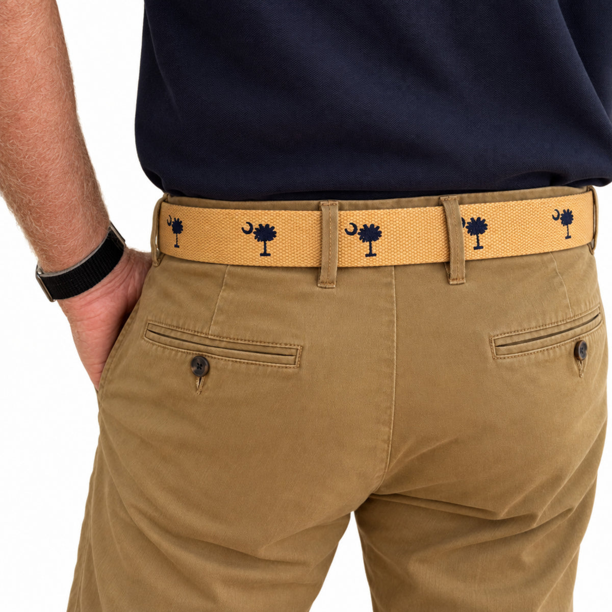 A close-up of a person wearing tan chinos with a mustard yellow woven belt featuring South Carolina flag belt  embroidered motifs, paired with a navy shirt.