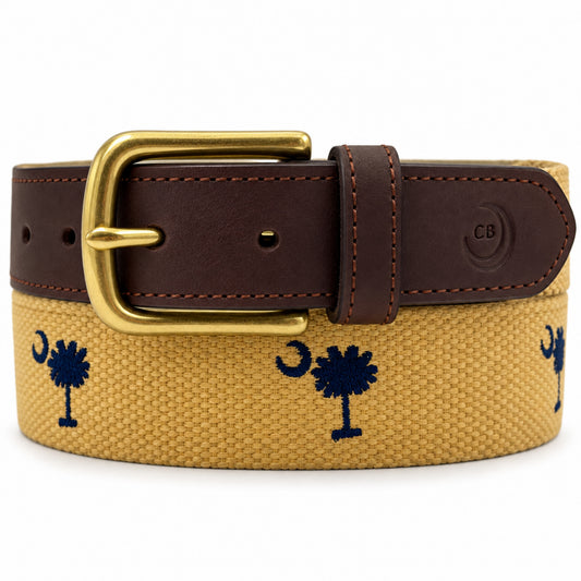 A coiled canvas belt with a dark brown leather strap, a gold buckle, and navy blue embroidered palm tree and crescent moon motifs on a tan canvas background. A circular 'CB' logo is embossed on the leather keeper. sc flag.