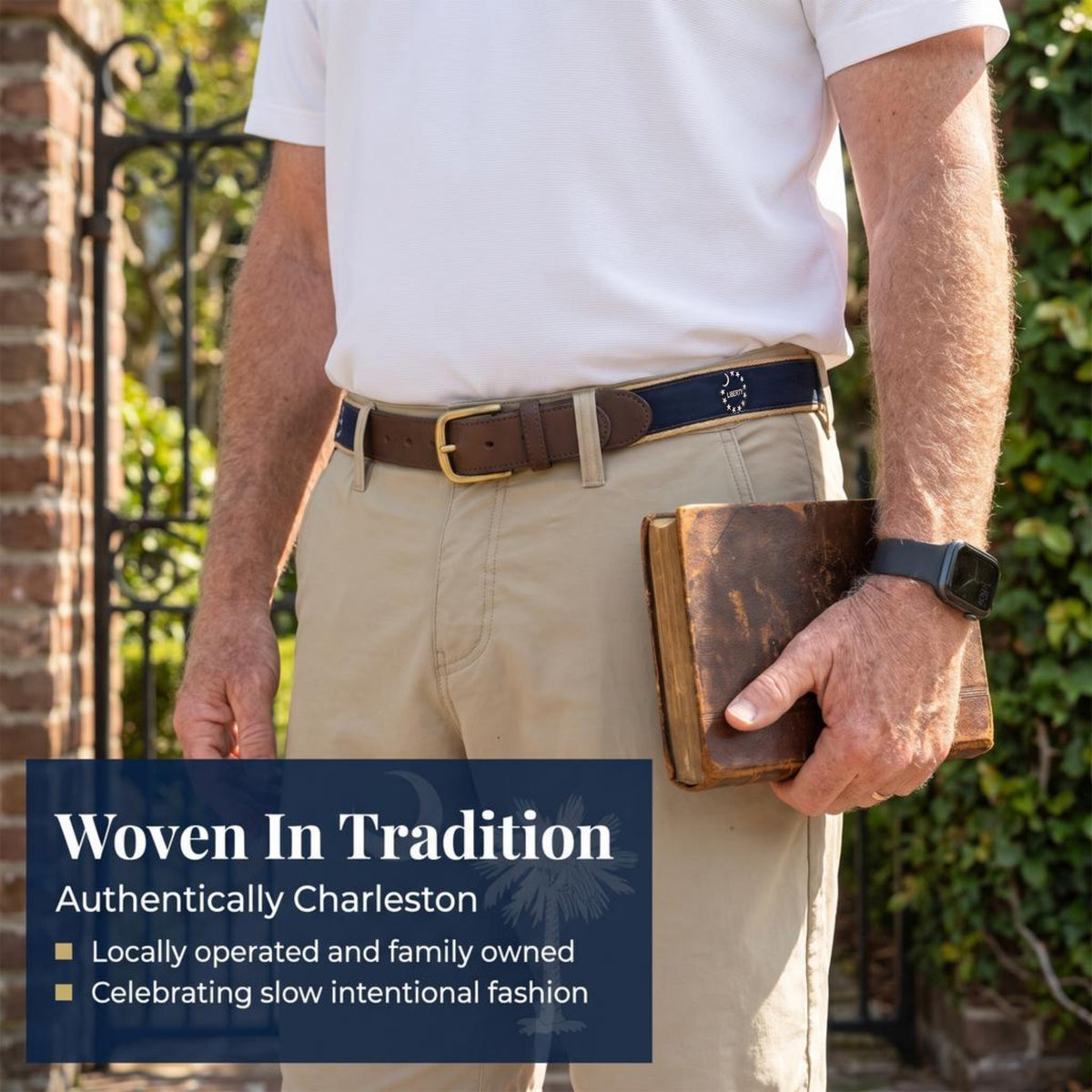 A man outdoors wearing a dark navy woven belt with a gold-tone buckle over a brown leather belt, paired with a white polo shirt and khaki pants. He is holding a brown leather book.