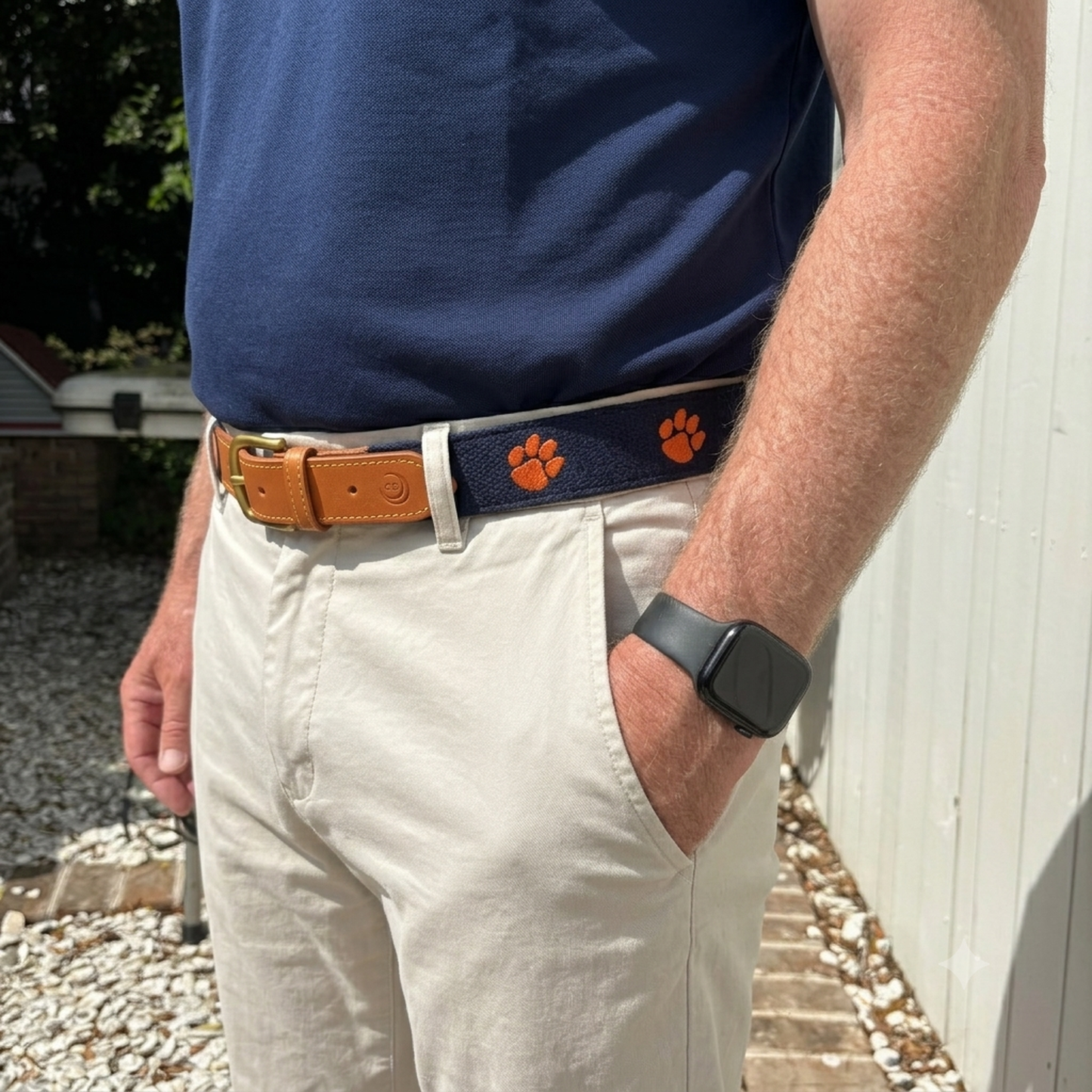 A close-up of a person wearing a navy blue belt with orange paw prints and a tan leather buckle, paired with beige pants and a navy shirt.  Clemson Tigers officially licensed logo and team name.