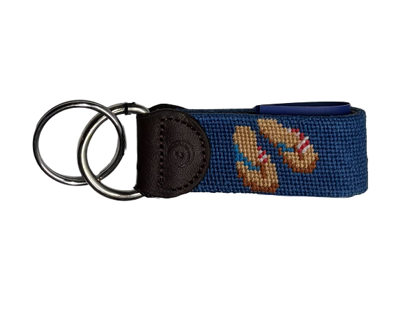 A navy blue hand-stitched needlepoint  keychain key fob featuring a embroidered pair of tan flip-flops with red and white and blue striped straps, attached to a dark brown leather tab and two stainless steel  metal key rings, shown on a plain white background.