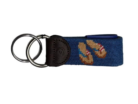 A navy blue hand-stitched needlepoint  keychain key fob featuring a embroidered pair of tan flip-flops with red and white and blue striped straps, attached to a dark brown leather tab and two stainless steel  metal key rings, shown on a plain white background.