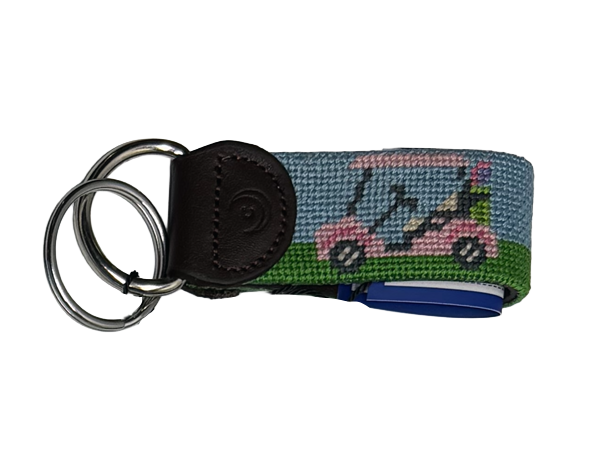 A golf-themed needlepoint key fob keychain featuring a pink green golf cart on a blue and green background, attached to a dark brown leather tabs and two stainless steel metal key rings.