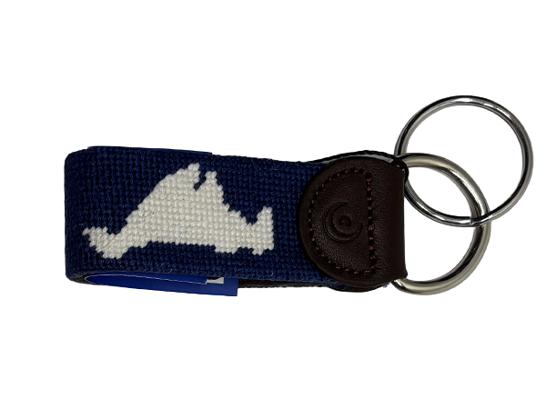 A navy blue woven keychain with a white embroidered Martha's Vineyard island  silhouette, a dark brown leather tab, and two silver metal key rings, shown on a plain white background.