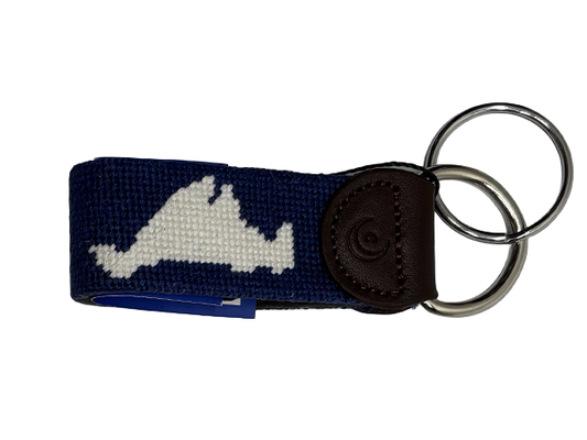 A navy blue woven keychain with a white embroidered Martha's Vineyard island  silhouette, a dark brown leather tab, and two silver metal key rings, shown on a plain white background.