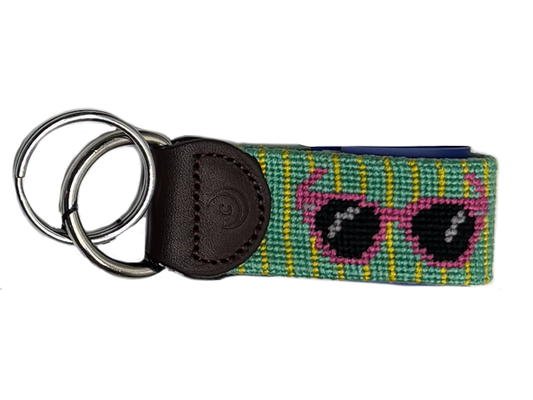 A woven keychain strap featuring a colorful pattern of green, yellow, and blue stripes with pink heart-shaped sunglasses, attached to a dark brown leather tab and two silver metal key rings, shown on a plain white background.